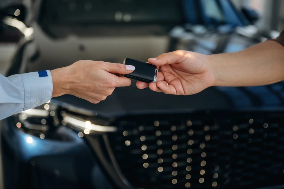 Une main en costume bleu marine transmet des clés de voiture à une main tendue, dans un environnement extérieur avec véhicule blanc en arrière-plan